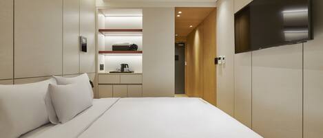 Een moderne hotelkamer met een groot bed, een flatscreen televisie aan de muur, een inbouwplank met een koffieapparaat en een houten deur die naar een andere kamer leidt.