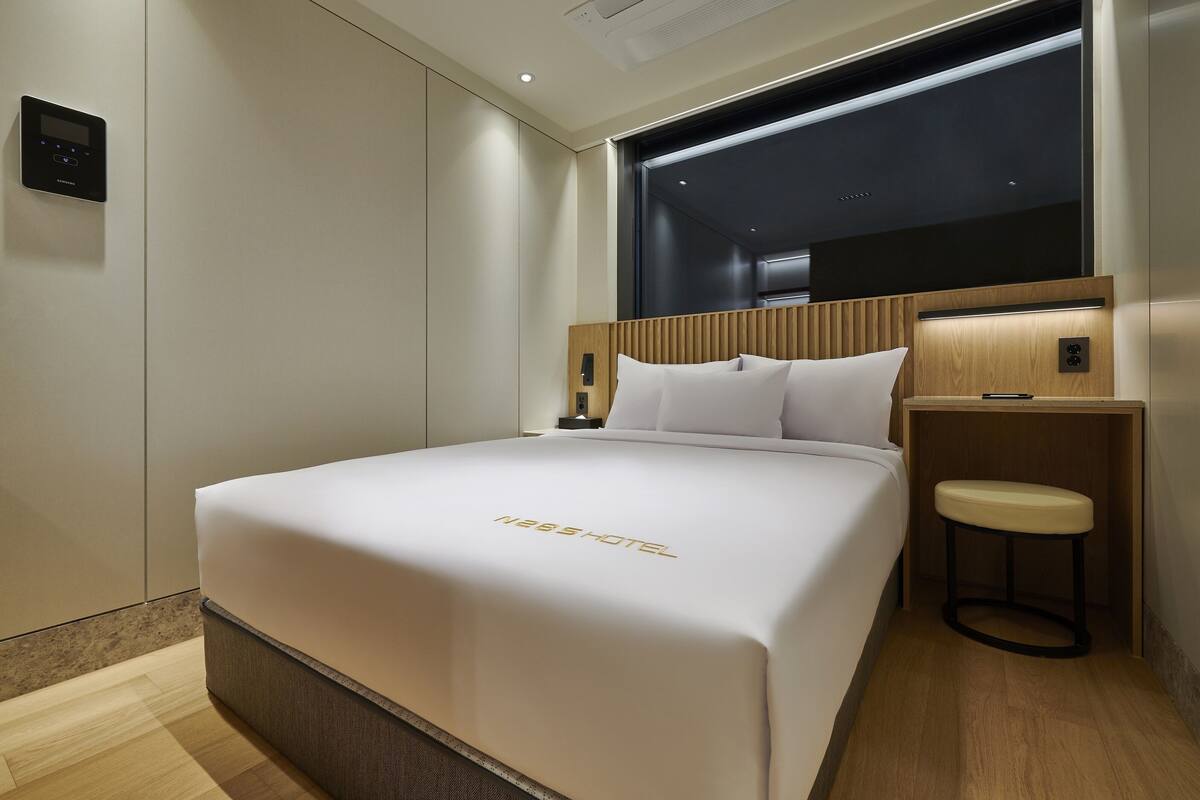 A hotel room with a large bed, a wooden headboard, a small stool, and a nightstand.