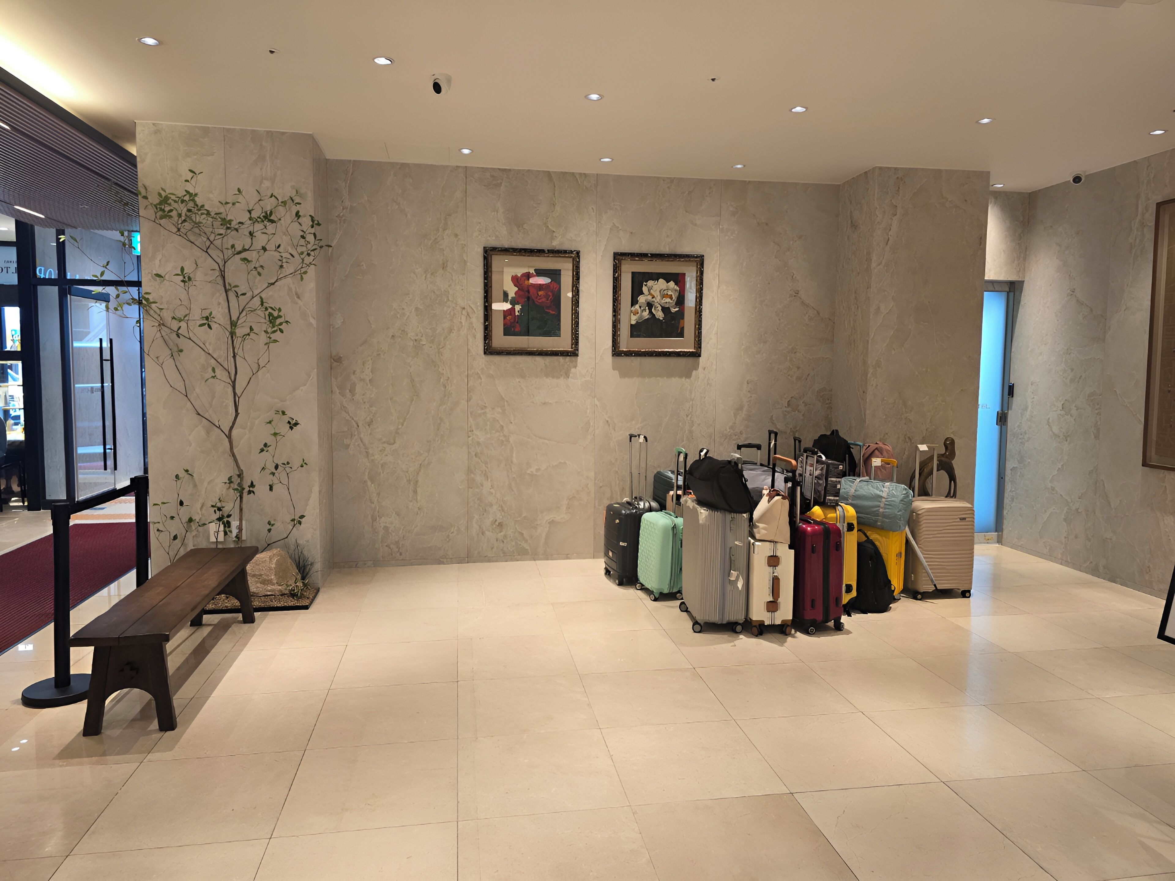 A spacious lobby with a marble floor, a wooden bench, a potted plant, and a row of suitcases.