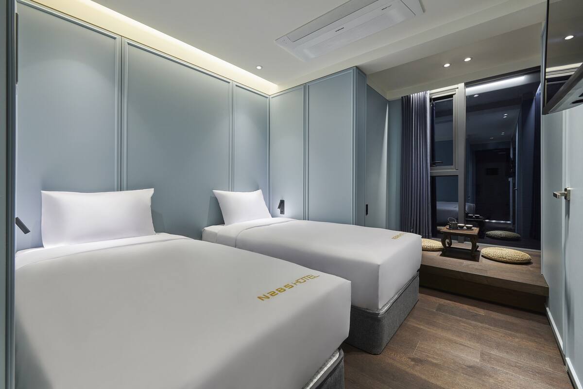 A hotel room with two beds, a small table with a lamp, and a view of the outside through a window.