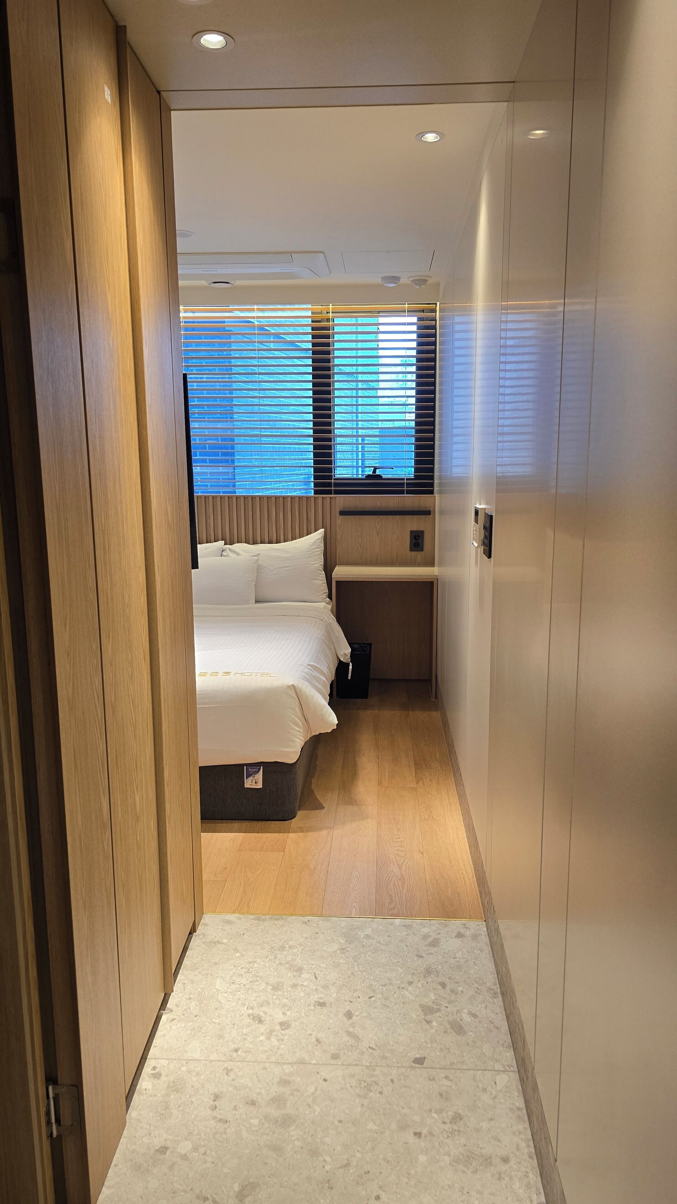 Un pasillo estrecho que conduce a un dormitorio con cama, un escritorio pequeño y una ventana con persianas.