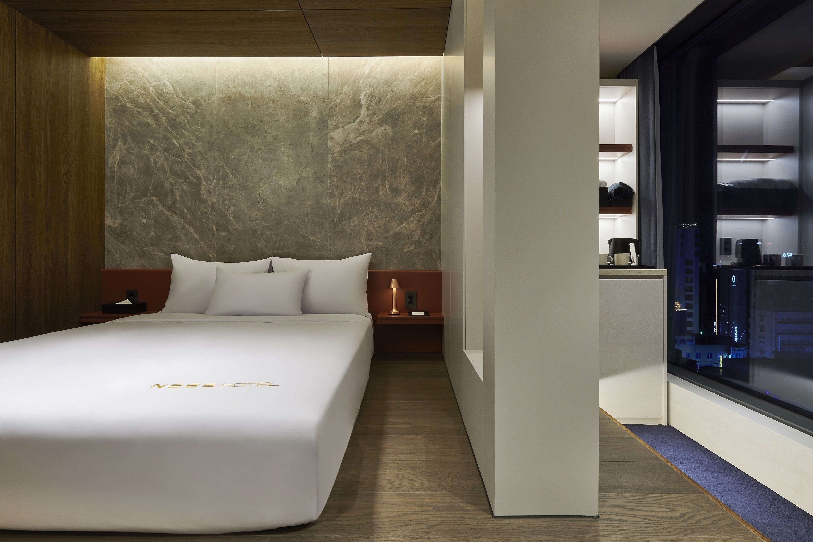 Una habitación de hotel moderna con una cama grande, una pared decorativa de mármol, una mesita de noche con lámpara y vistas a la ciudad a través de una ventana.