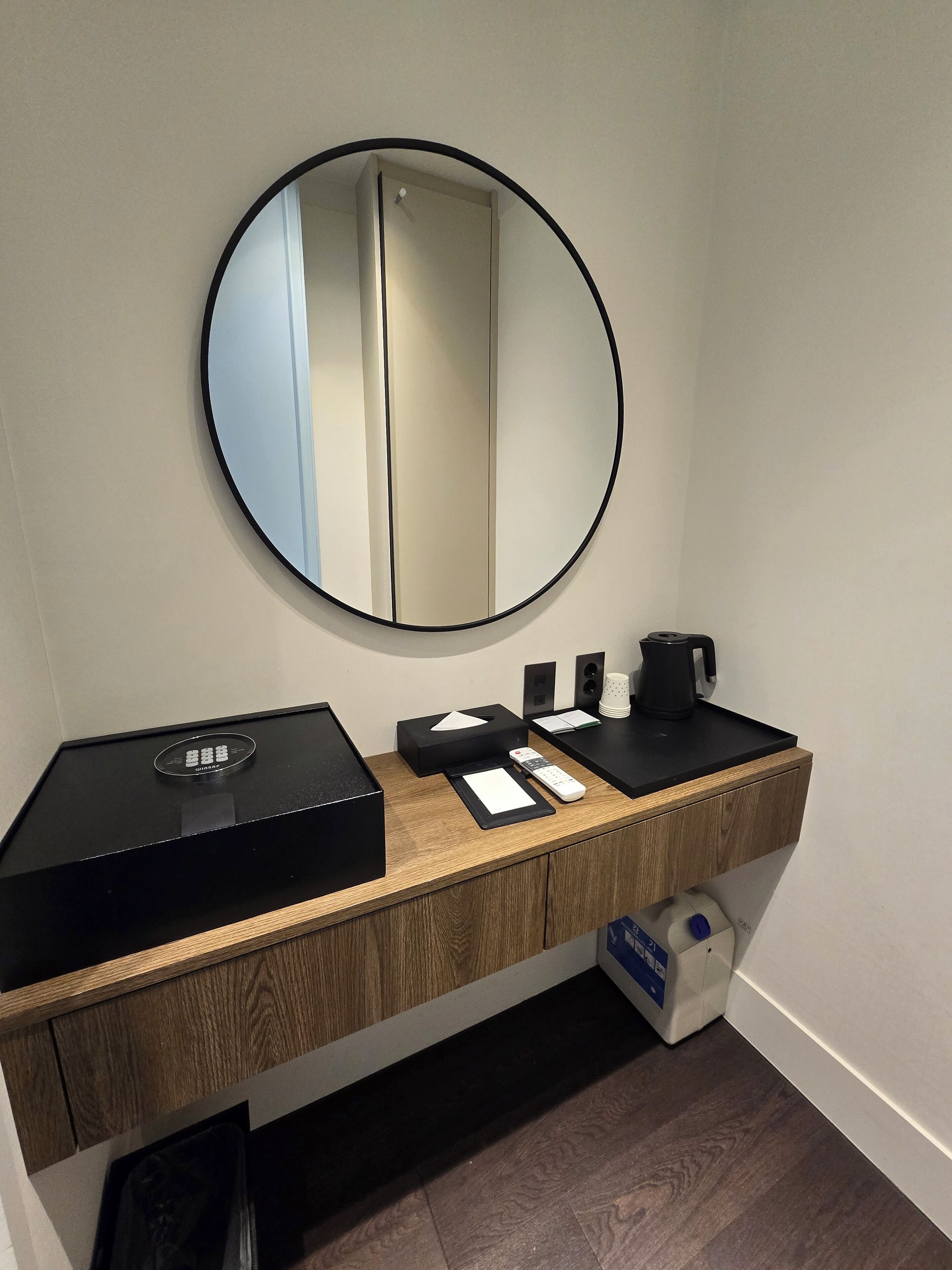 A modern bathroom with a wooden vanity, a round mirror, a black electric kettle, a remote control, and a tissue box.