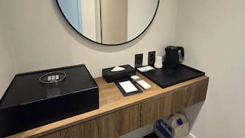 A modern bathroom with a wooden vanity, a round mirror, a black electric kettle, a remote control, and a tissue box.