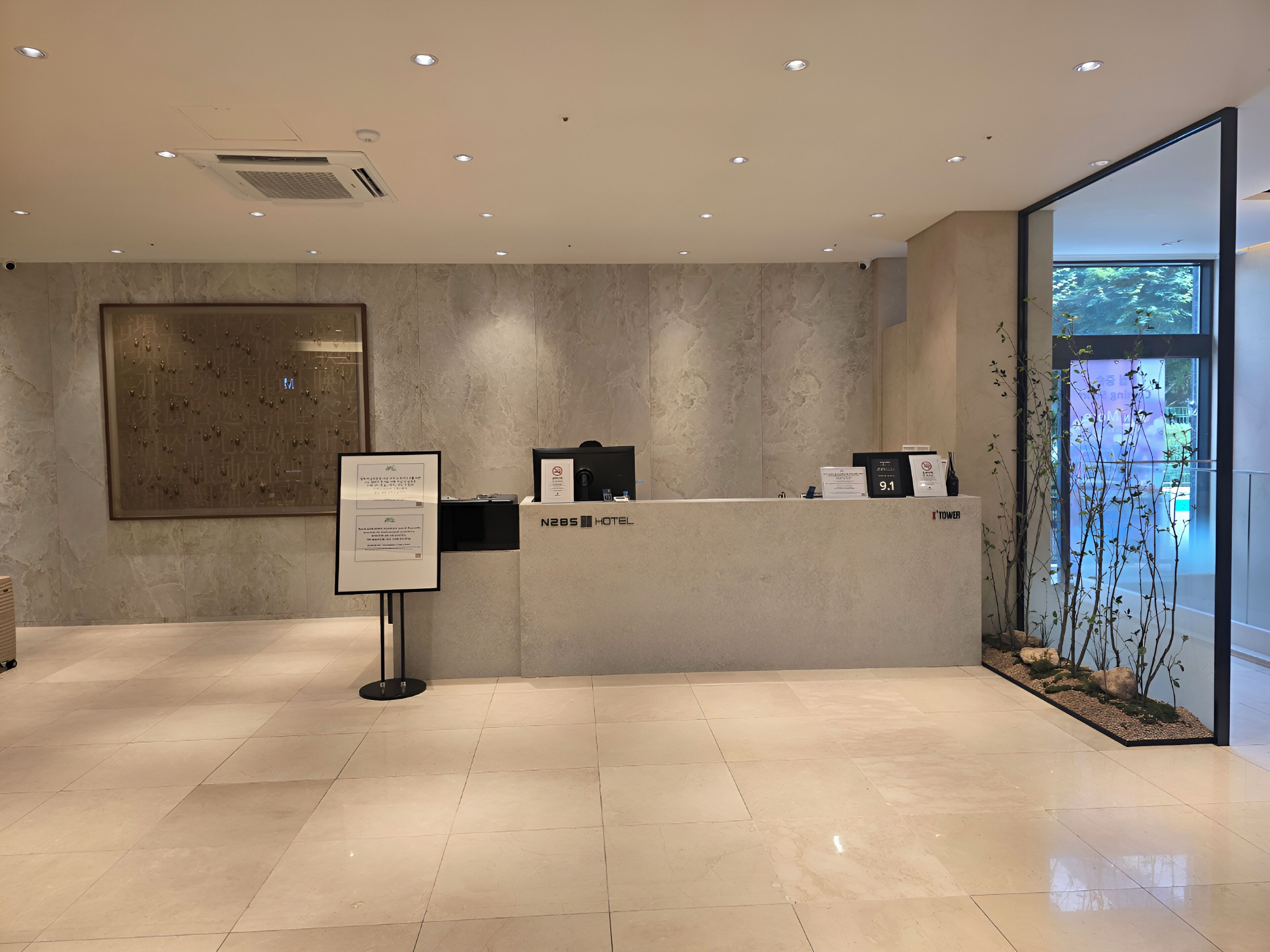 A hotel reception area with a modern design, featuring a reception desk, a large framed artwork on the wall, and a view of the outdoors through a glass door.