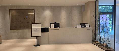 A hotel reception area with a modern design, featuring a reception desk, a large framed artwork on the wall, and a view of the outdoors through a glass door.
