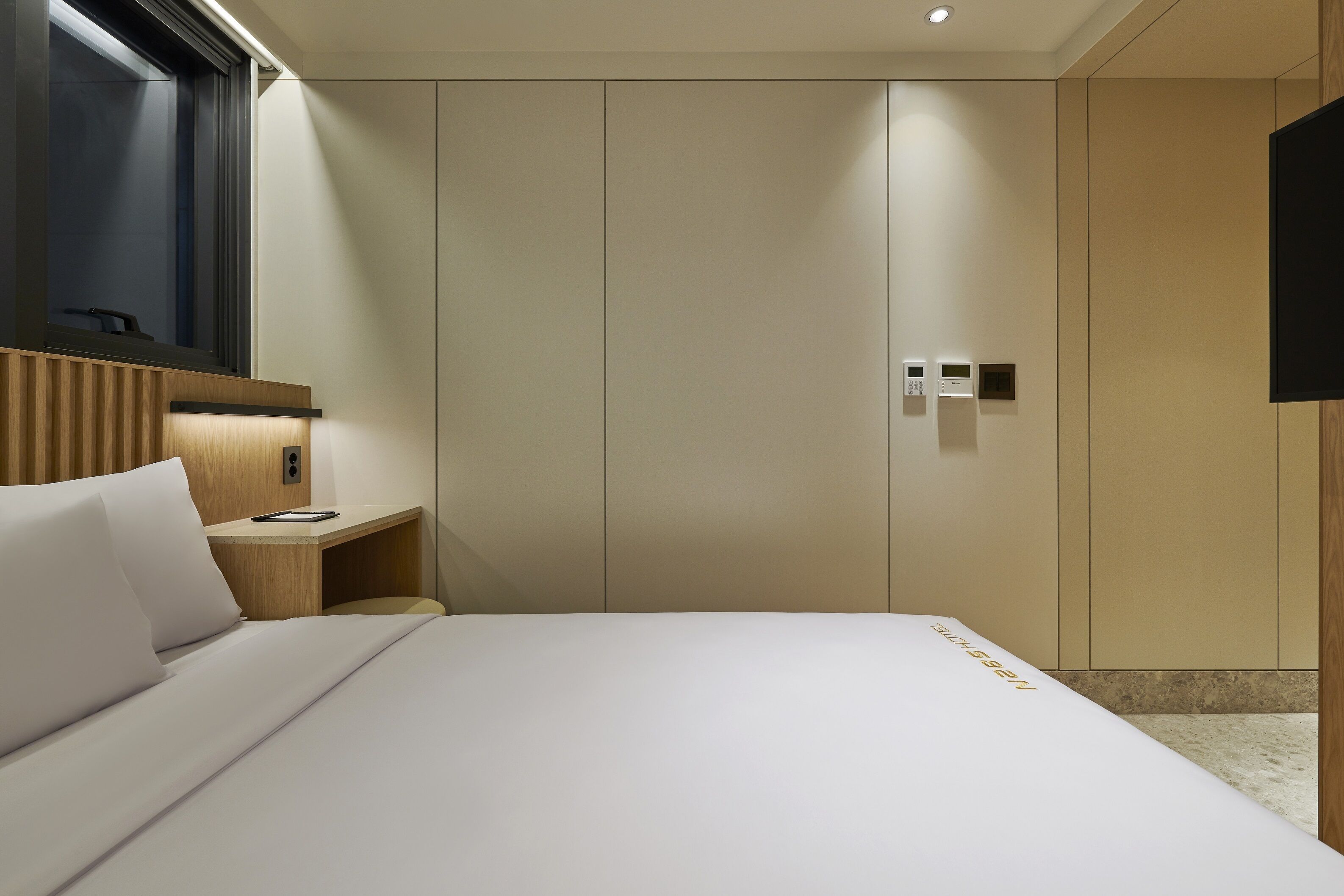 A hotel room with a bed, a bedside table, a TV, and a control panel on the wall.