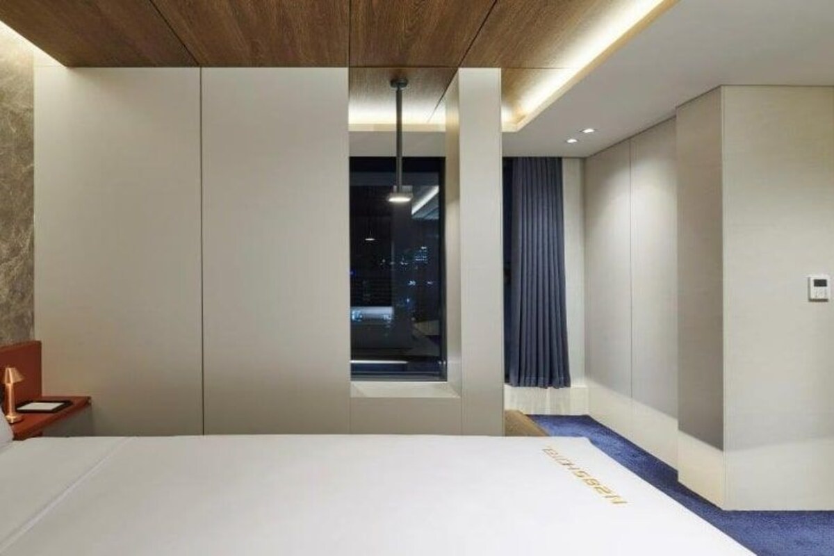 Una habitación de hotel moderna con una cama grande, una mesita de noche, un ventanal con vista a la ciudad iluminada y un pequeño balcón.