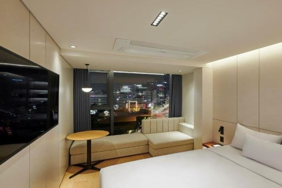 A modern hotel room with a large bed, a seating area with a small table, a city view through large windows, and a flat-screen TV mounted on the wall.