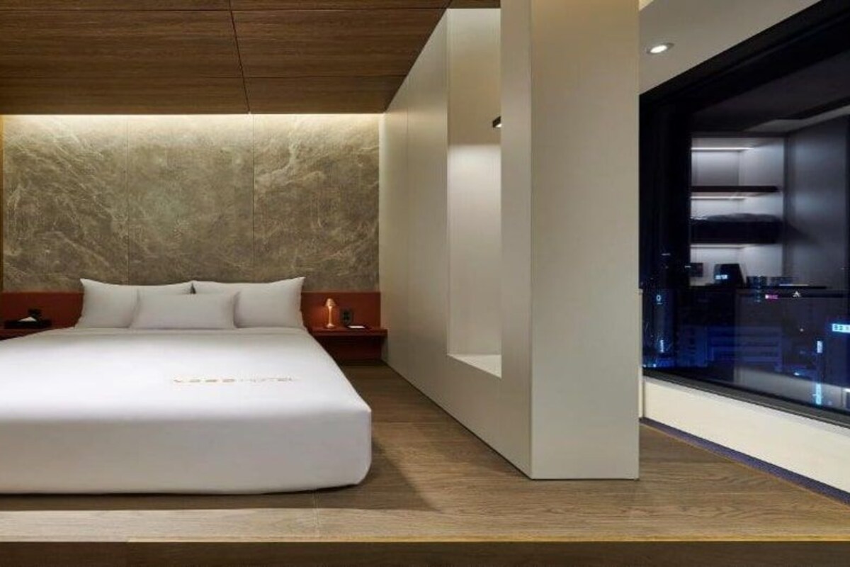 A modern bedroom with a large bed, wooden paneling on one wall, and a view of the city through a window.