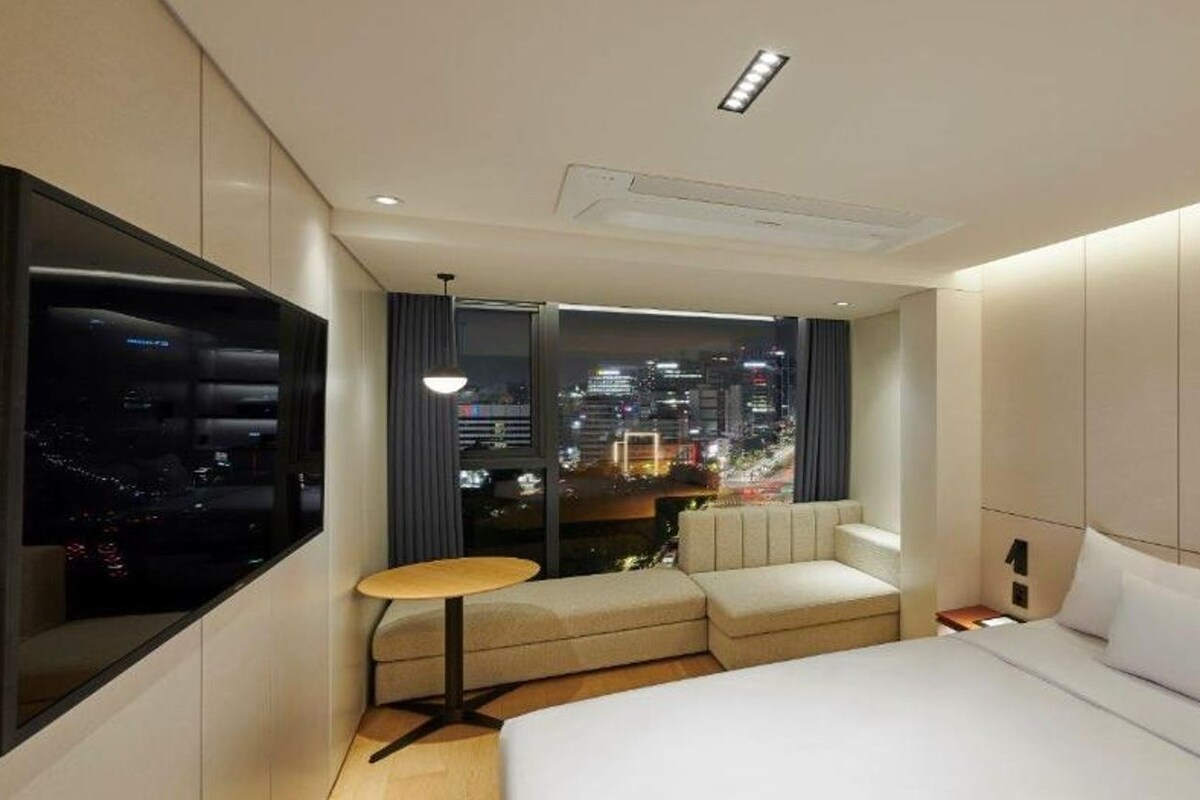 Una habitación de hotel moderna con una cama grande, una zona de estar con una pequeña mesa y vistas a la ciudad a través de amplios ventanales.