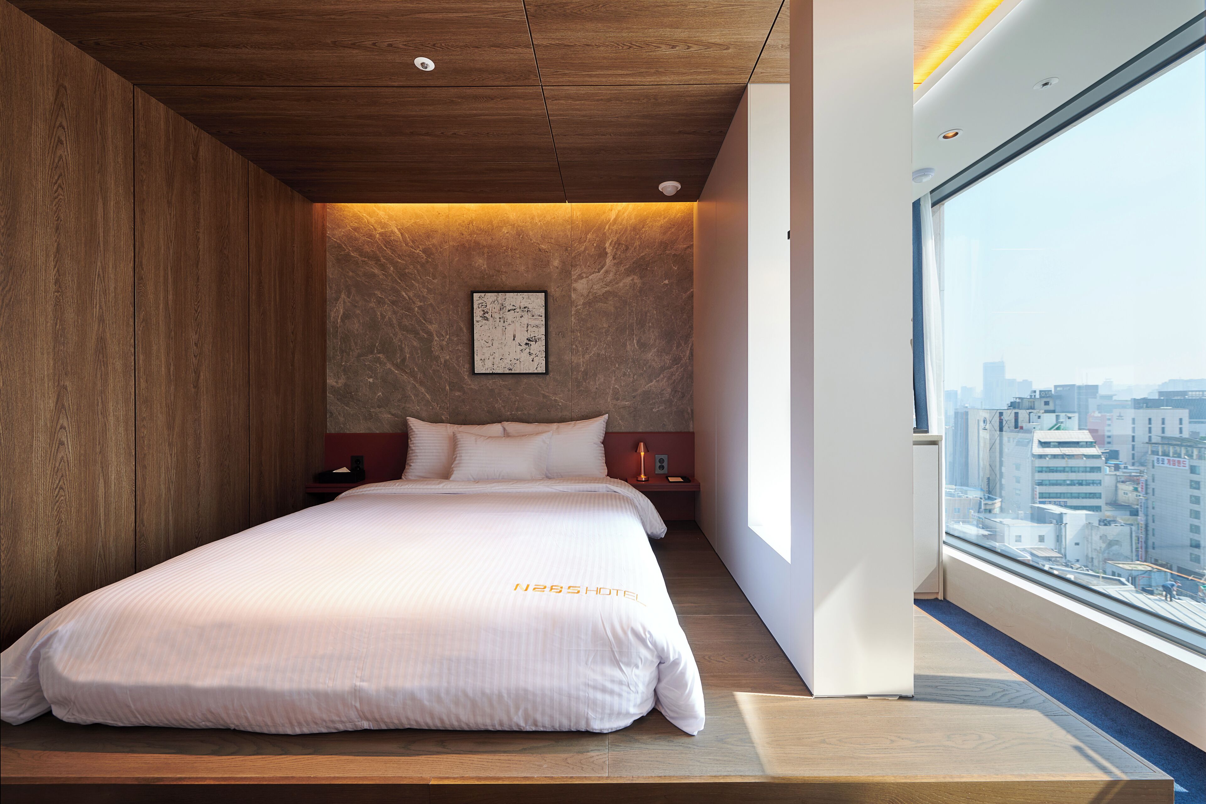 Una habitación de hotel moderna con una cama grande, techo de madera y vista a la ciudad a través de un ventanal amplio.