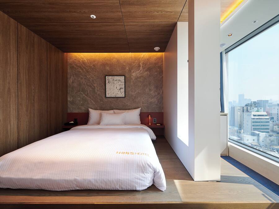 Una habitación de hotel moderna con una cama grande, techo de madera y vista a la ciudad a través de un ventanal amplio.