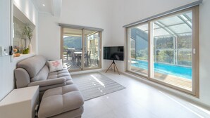 A modern living room with a sofa, a TV on a tripod, and large sliding glass doors leading to a pool. - Gapyeong Jinikidsdogchaepoolvilla (Gapyeong)