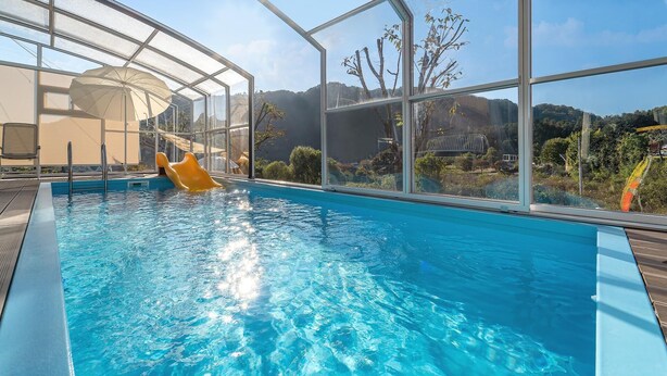 An indoor swimming pool with clear blue water, a yellow slide, and a glass enclosure with a view of the surrounding landscape. - Gapyeong Jinikidsdogchaepoolvilla (Gapyeong)