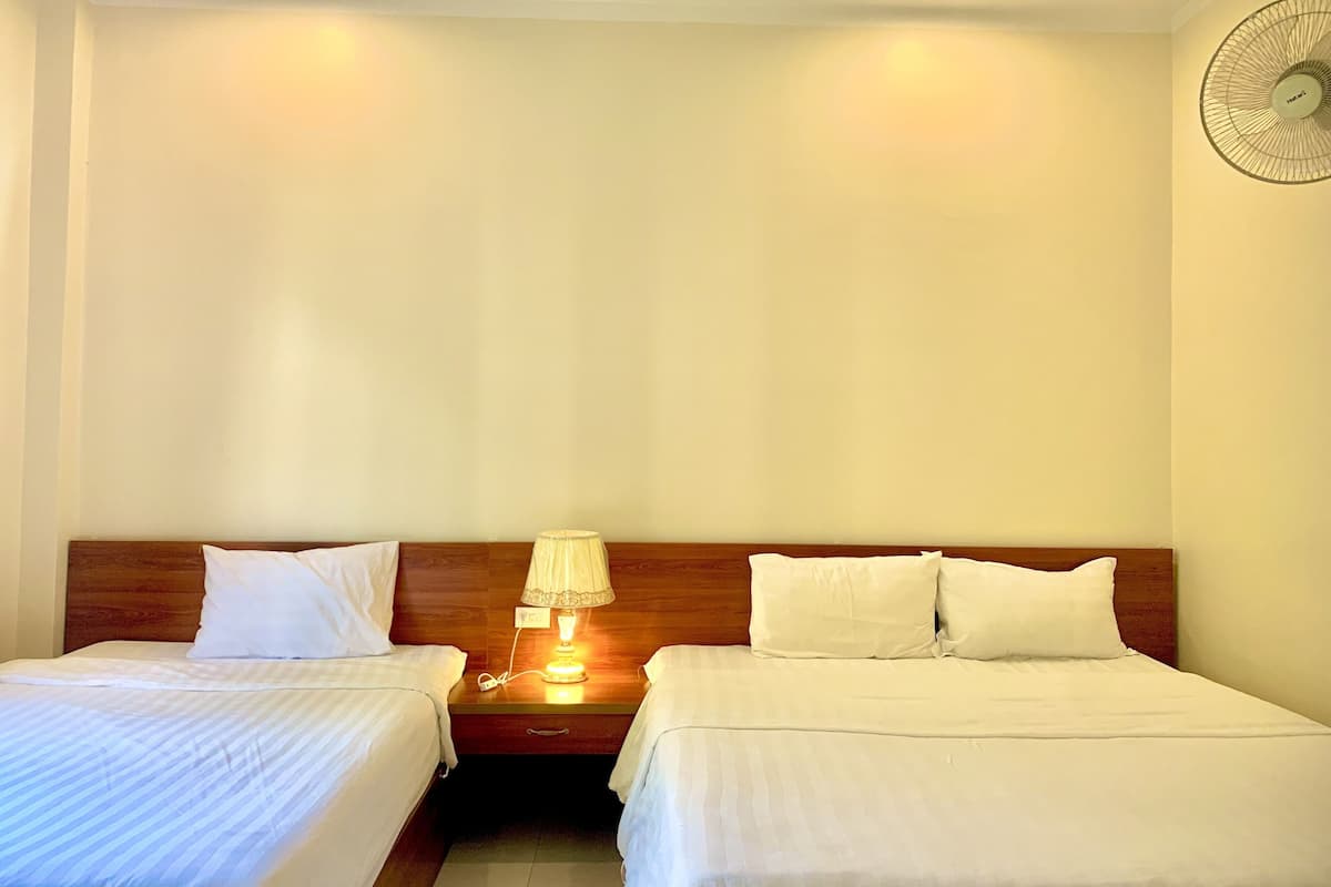 Una camera d'albergo con due letti singoli, un comodino con una lampada e una ventola a muro.