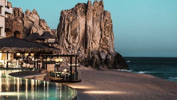A beachfront hotel with a pool, a thatched-roof bar area, and a rocky cliff backdrop. - Luxury oceanfront resort suite with access to Greg Norman Signature golf course! (Cabo San Lucas)