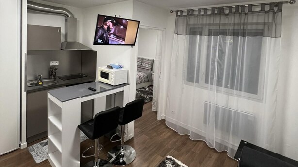 Un espace de vie compact comprenant un coin cuisine, un espace repas avec des tabourets de bar, un téléviseur fixé au-dessus du comptoir et une grande fenêtre agrémentée de rideaux légers.