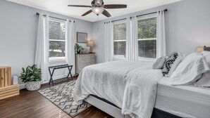 Une chambre à coucher comprenant un lit, un ventilateur de plafond, des fenêtres avec stores, une table de chevet, une lampe, une plante et un banc en bois.