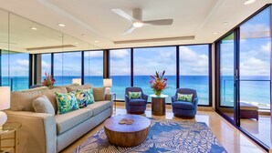 A modern living room with a large sofa, two armchairs, a coffee table, and a view of the ocean through large windows.