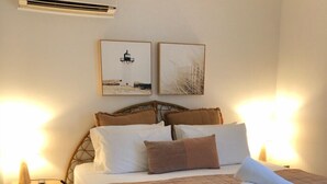 Un lit bien fait, avec une couverture marron, deux oreillers et une serviette enroulée, posé sur un sommier de couleur foncée. On y trouve deux tables de chevet avec des lampes, et un climatiseur mural fixé au-dessus du lit.