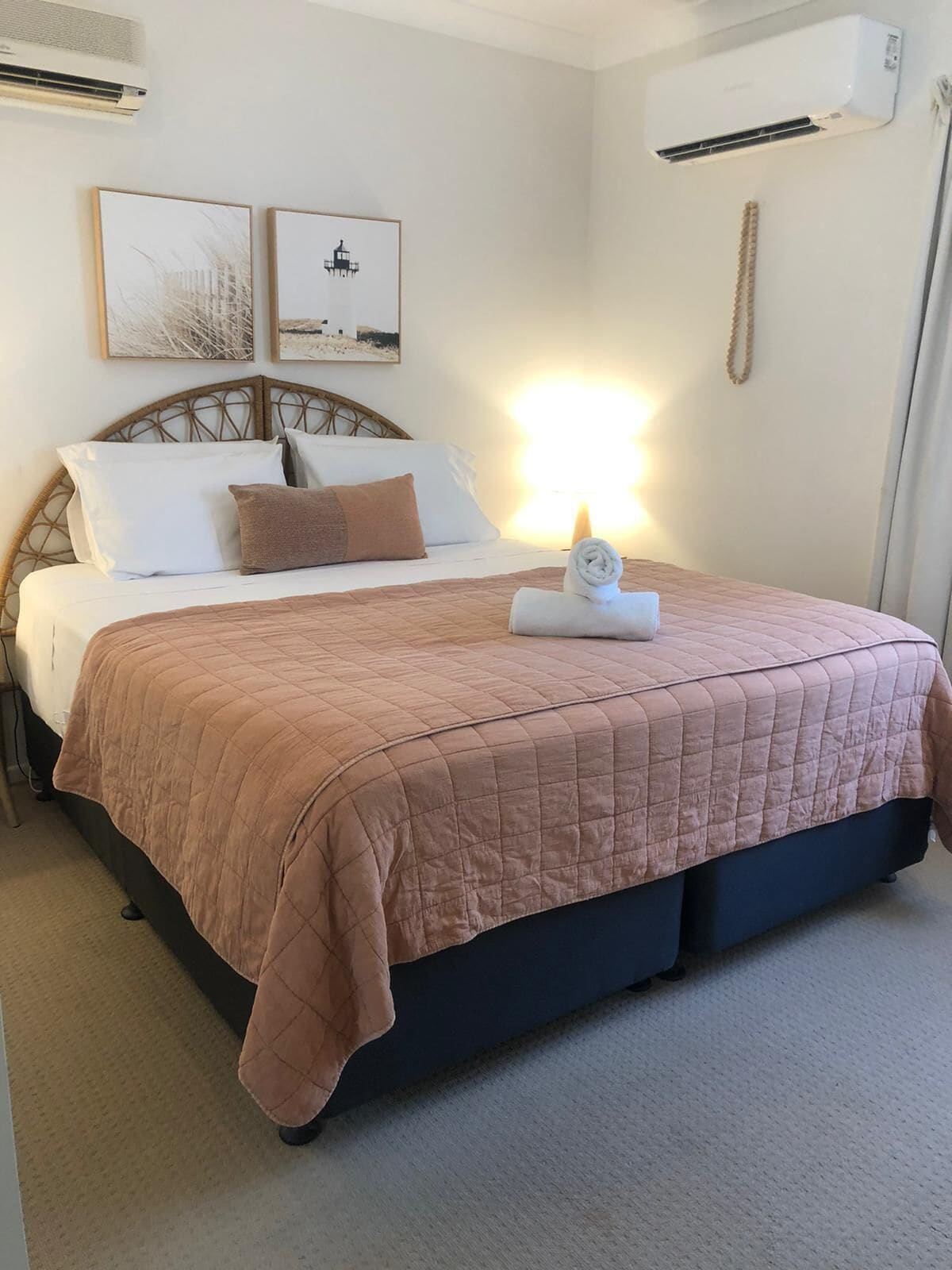 Una cama bien hecha con un edredón rosa, una almohada blanca y un plaid marrón. Se observa un aire acondicionado de pared y dos cuadros enmarcados sobre la cama.