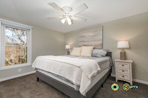 A bedroom with a bed, a ceiling fan, two bedside tables with lamps, a window with a view of trees, and a wall-mounted artwork. - Cozy Renovated Woodland Townhome | Minutes from Bryce Resort! (Basye)
