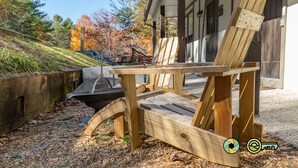 A wooden Adirondack chair with a metal frame, placed on a gravel surface near a building with a porch.