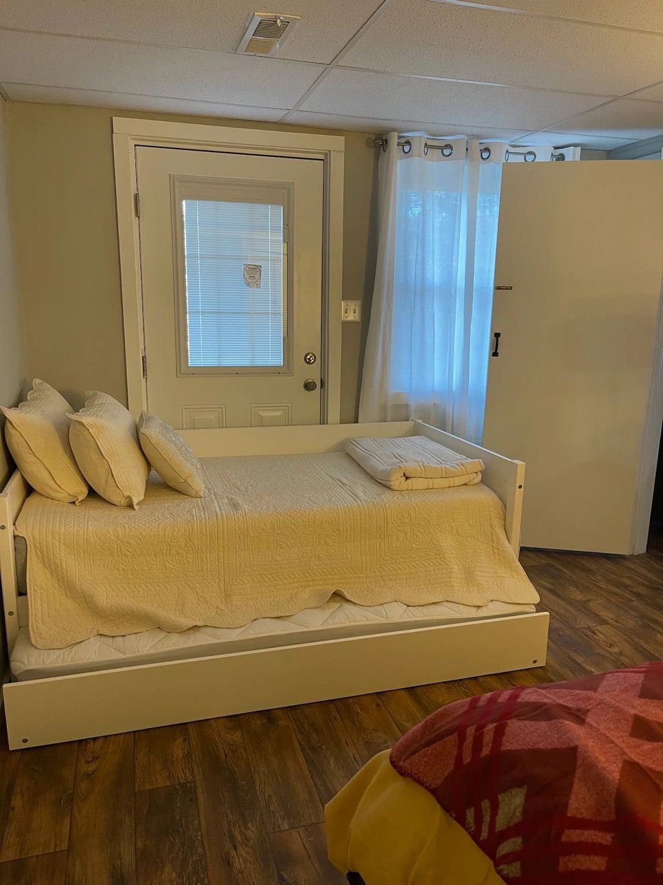 A single-bed room with a door, a window with blinds, a curtain, and a wooden floor.