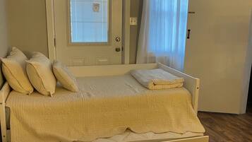 A single-bed room with a door, a window with blinds, a curtain, and a wooden floor.