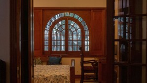 A bedroom with a large arched window, a bed with floral bedding, a wooden chair, and a table with a decorative object.