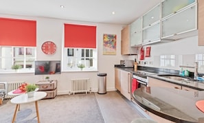 A modern kitchen with a large island, red window blinds, a clock, and a colorful painting.