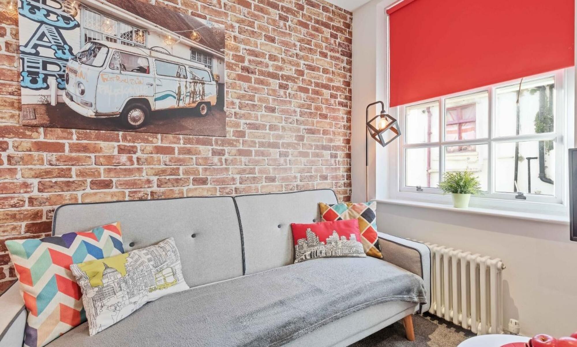 Une pièce chaleureuse avec un mur en briques, une fresque représentant un vieux van, un canapé gris agrémenté de coussins colorés, un store rouge et un radiateur.
