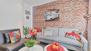 A living room with a brick wall, a grey sofa, colorful cushions, a small table with fruit, and a plant. - Majestic Mews Apartment Super Central Sleeps 2 to 8 Guests Free Wifi (Brighton)