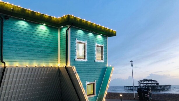 A teal-colored building with two windows and a slanted roof, situated by a beach with a pier in the background. - Majestic Mews Apartment Super Central Sleeps 2 to 8 Guests Free Wifi (Brighton)