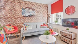 A cozy living room with a brick wall, a grey sofa, a red chair, a round table with a plant, and a TV on the wall. - Majestic Mews Apartment Super Central Sleeps 2 to 8 Guests Free Wifi (Brighton)