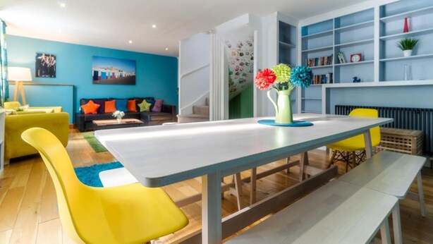 A modern living room with a dining table, yellow chairs, a sofa with colorful cushions, and a bookshelf. - Pebble Mews House - Sleeps 6 to 8 Guests (Brighton)