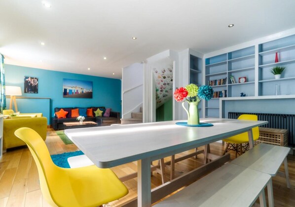 Una sala de estar moderna con una mesa de comedor, sillas amarillas, un sofá con cojines de colores y una estantería.