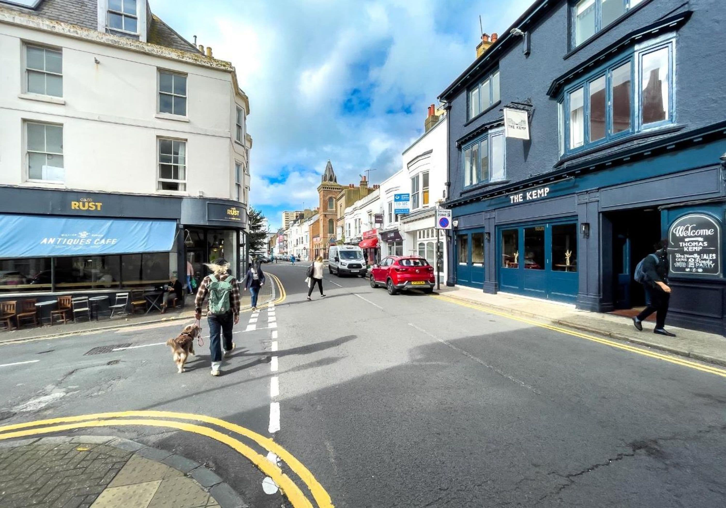 A street view with buildings, a red car parked on the side, and pedestrians.