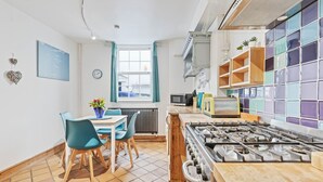 A modern kitchen with a gas stove, a dining table with chairs, a window with curtains, and a tiled backsplash. - Pebble Mews House - Sleeps 6 to 8 Guests (Brighton)