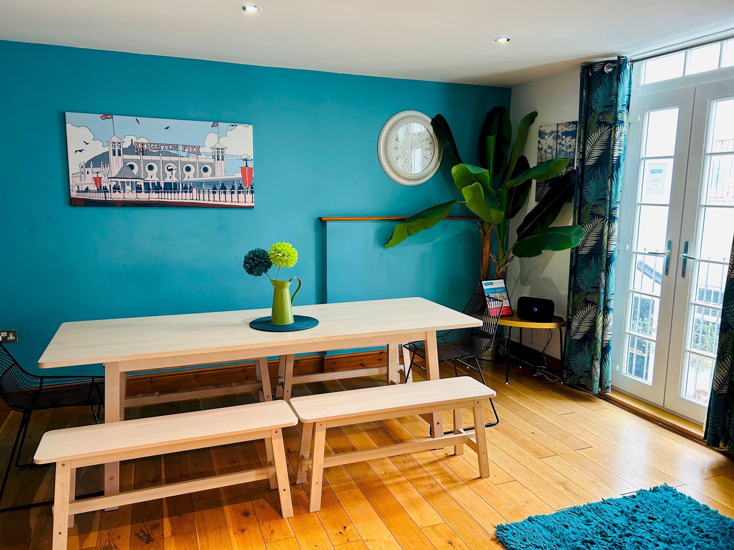 A room with a blue wall, a wooden table with benches, a clock, a large plant, and a window with curtains.