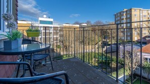 A balcony with a round table and chairs, a potted plant, and a view of surrounding buildings and greenery.
