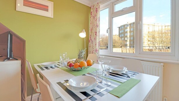 A dining area with a table set for two, a fruit bowl, and a view of buildings through the window.