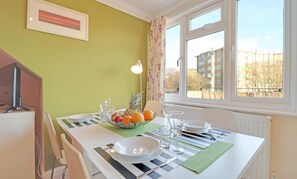 Spiseplads med et bord dækket til to, en skål med frugt og udsigt til bygninger gennem vinduet.