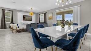 Ein moderner Essbereich mit einem weißen Tisch und blauen Samtstühlen, einem grauen Sofa und einem großen Fenster mit Blick.