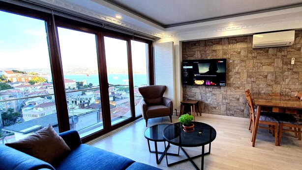 Een moderne woonkamer met een groot raam, een flatscreen televisie aan een stenen muur, een eethoek met houten meubels en uitzicht op de stad en zee.