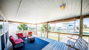 A covered outdoor area with a wooden deck, red chairs, a small table with a vase, and a swing. - Bounty Pet Friendly · Entertain & Relax at our Beach Home - Pet Friendly (Galveston)