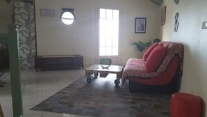 A living room with a sofa, a coffee table, and a red ottoman. There is a window with blinds, a radiator, and a plant on the wall. - Le Gosier Spacious loft with small sea view (Le Gosier)