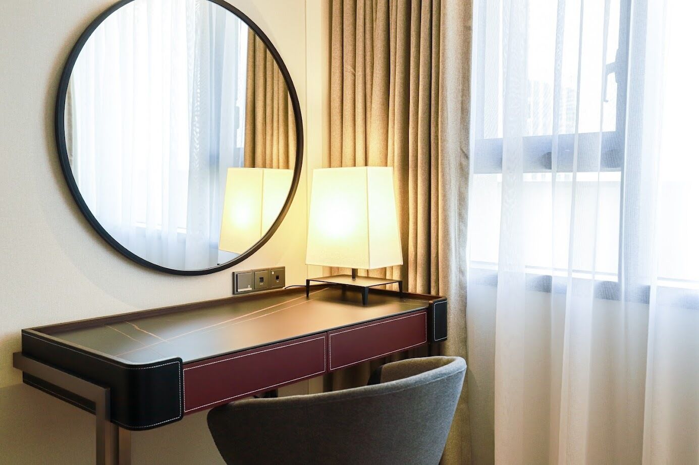 A hotel room with a desk, a chair, a round mirror, a lamp, and a window with curtains.
