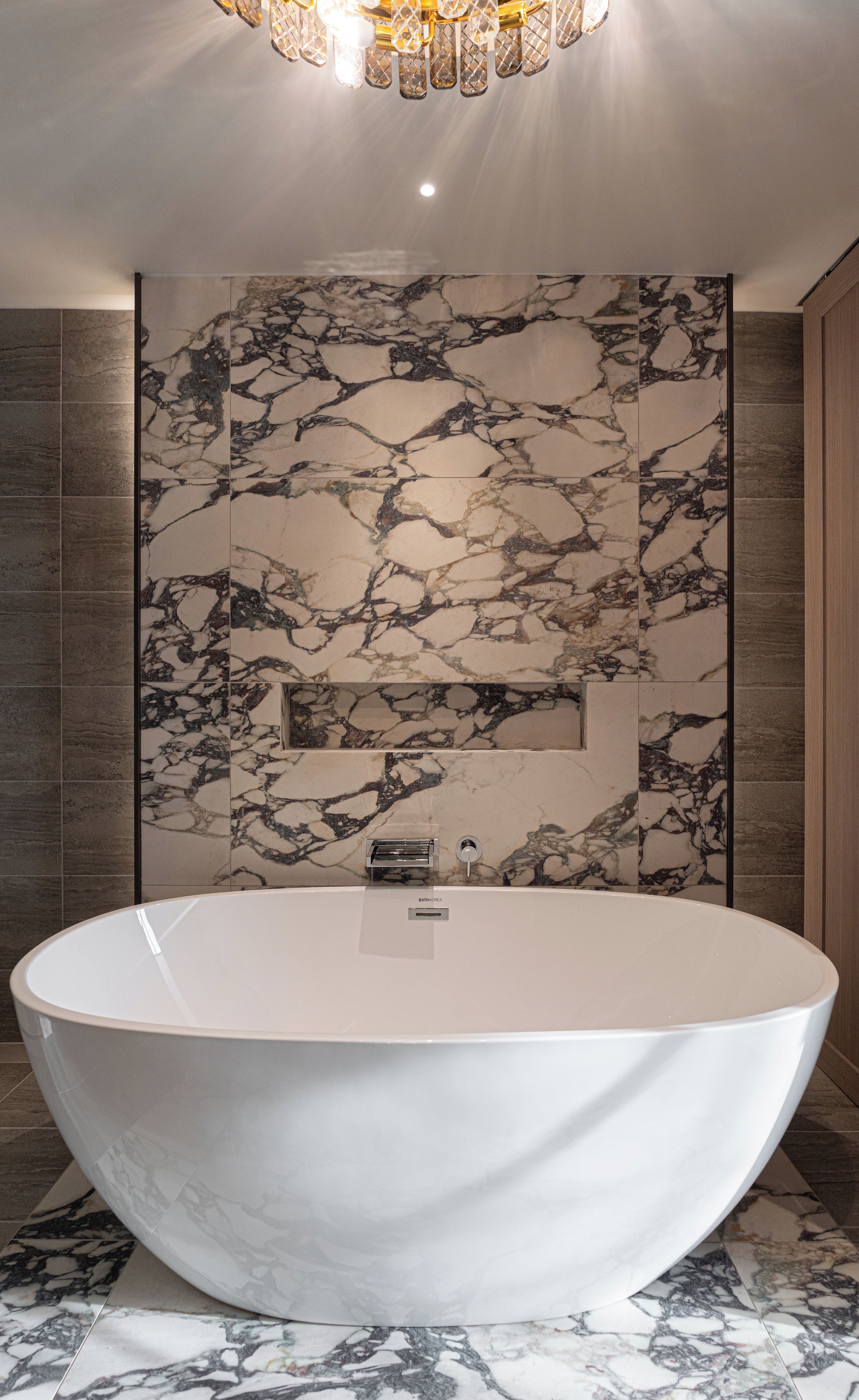 A modern bathroom with a freestanding bathtub, marble wall tiles, and a chandelier.